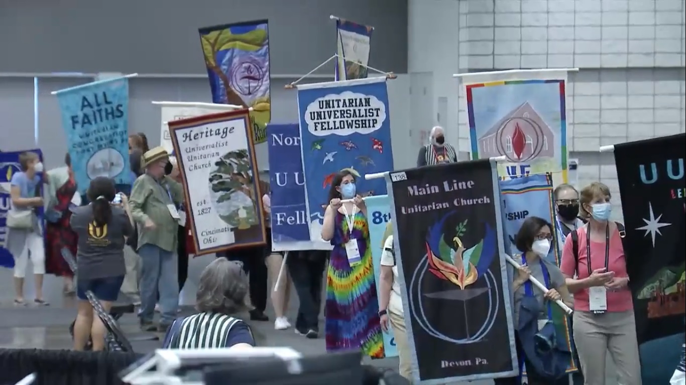People of various ages in COVID masks gather in a rough line holding vertical banners on poles, each banner representing a different Unitarian Universalist church.