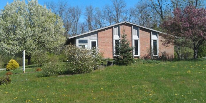 A one-story brick building with vertical windows sits on green grass surrounded by trees.