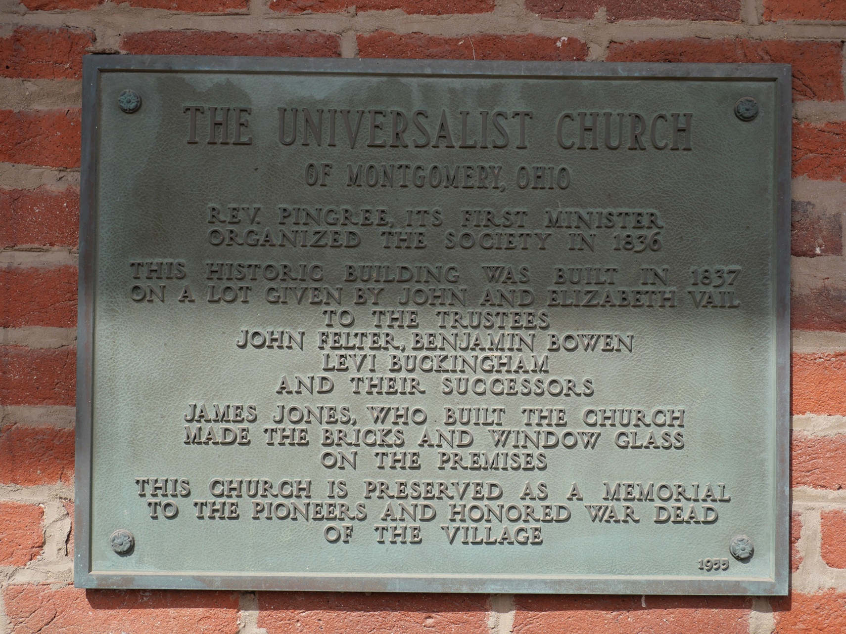 A metal plaque on an old brick wall. The plaque reads, in part: "THE UNIVERSALIST CHURCH of Montgomery, Ohio. Rev. Pingree, its first minister, organized the society in 1836. This historic building was built in 1837.... James Jones, who built the church, made the bricks and window glass on the premises. This church is preserved as a memorial to the pioneers and honored war dead of the village."