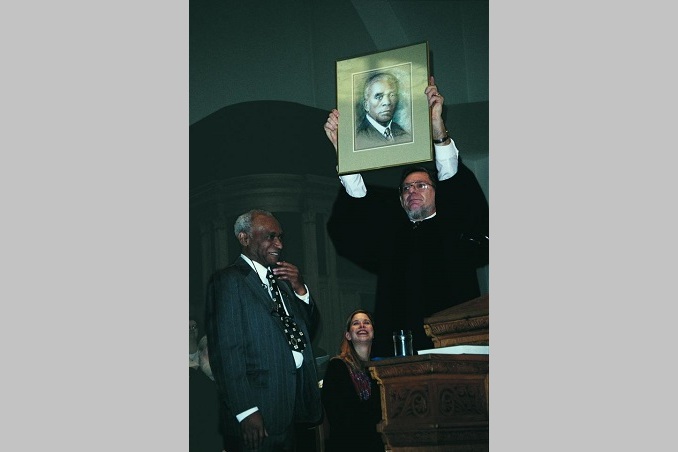 At a pulpit, a male minister in robes holds up a photograph of WHG Carter, an African American minister in the early 20th century. Rev. Carter's grandson stands and looks on. Another minister, seated, looks on.